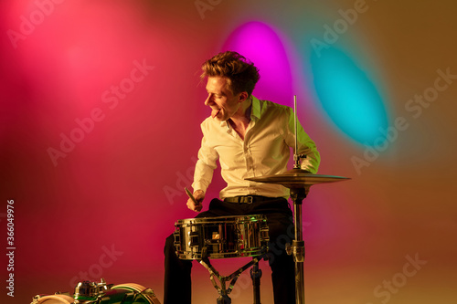 Cheerful. Young caucasian inspired and expressive musician, drummer performing on multicolored background in neon. Concept of music, hobby, festival, art. Joyful artist, colorful, bright portrait.