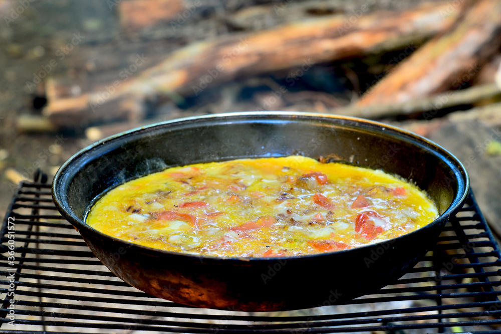 Fried eggs with vegetables are cooked in a pan on a grill on coals in the open air.