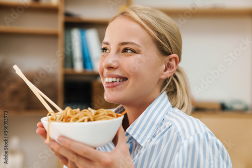 Image of cheerful blonde woman eating noodle while working in office