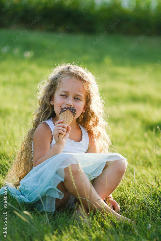Little blonde girl with long hair has fun eating ice cream in a summer park. Little blonde girl with long hair has fun eating ice cream in a summer park. Sits on the grass with ice cream. Little blonde girl with long hair has fun eating ice cream in a summer park. Little blonde girl with long hair has fun eating ice cream in a summer park. Sits on the grass with ice cream.