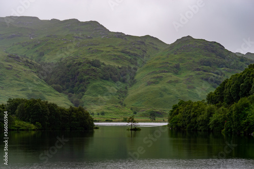 A lonely tree on a lake surrounded by water with mountain ridge building up behind it during a hazy day.
