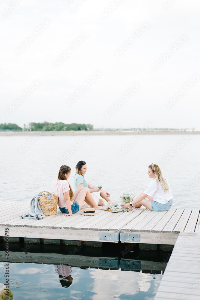 three girls sitting on a wooden pier in front of the water. rest, picnic,lemonade