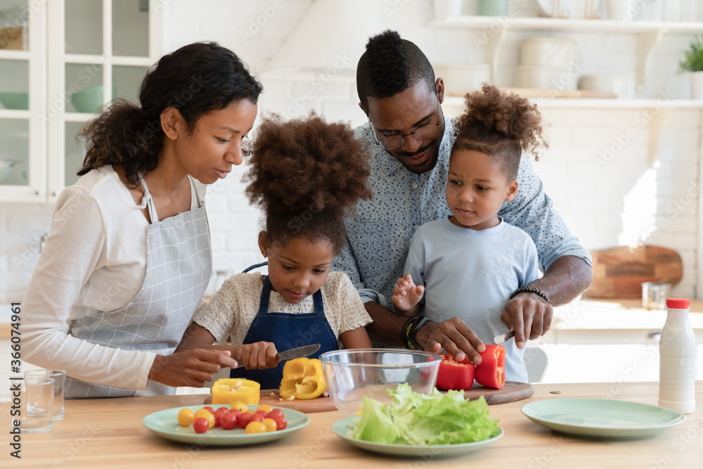 Loving african American mom and dad teach cooking chop vegetables with excited little children in kitchen, happy caring biracial parents prepare food organic salad with small ethnic kids at home
