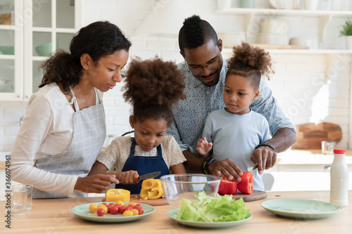 Loving african American mom and dad teach cooking chop vegetables with excited little children in kitchen  happy caring biracial parents prepare food organic salad with small ethnic kids at home