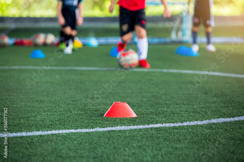 selective focus to red cone marker is soccer training equipment on green artificial turf with blurry kid players training background. Material for trainning class of football academy.