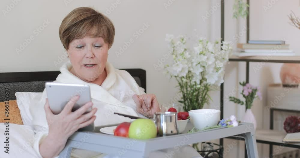 Serious old lady in rob talking during video call while having breakfast in bed. Woman pensioner communication on tablet using frontal camera while sitting with trey full of food. Serious old lady in rob talking during video call while having breakfast in bed. Woman pensioner communication on tablet using frontal camera while sitting with trey full of food.