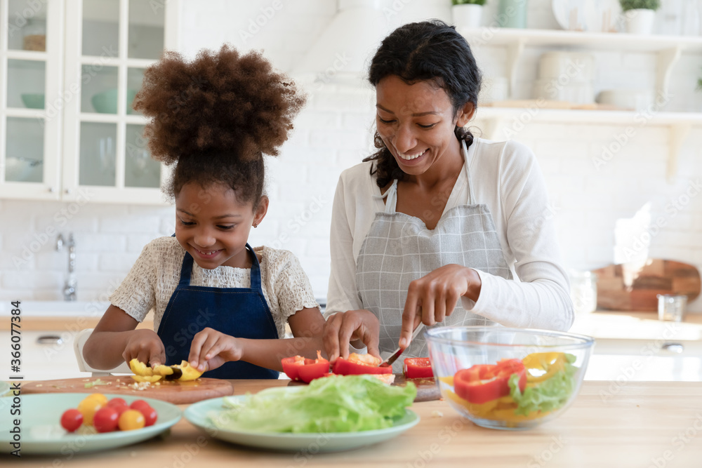 Smiling young african American mother and small daughter chop vegetables prepare healthy vegetarian salad in kitchen, happy biracial mom teach little girl child cooking food at home, dieting concept