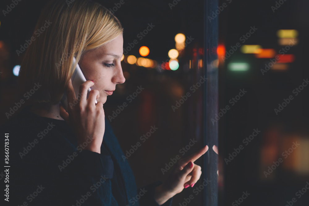 Obraz premium Young woman speaking on mobile phone while using automated teller machine with big digital screen which reflect beautiful light on her face, female verifies account balance on banking application