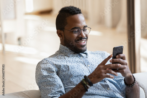 Smiling young african American man in glasses sit relax on couch at home browse wireless internet on modern cellphone, happy biracial male in spectacles look at screen message text use smartphone