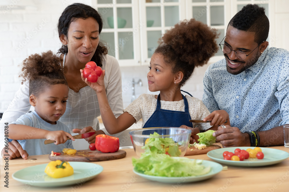Happy young african American family with two little kids have fun cooking together at home kitchen, loving smiling biracial parents teach small children prepare healthy food organic salad or dinner