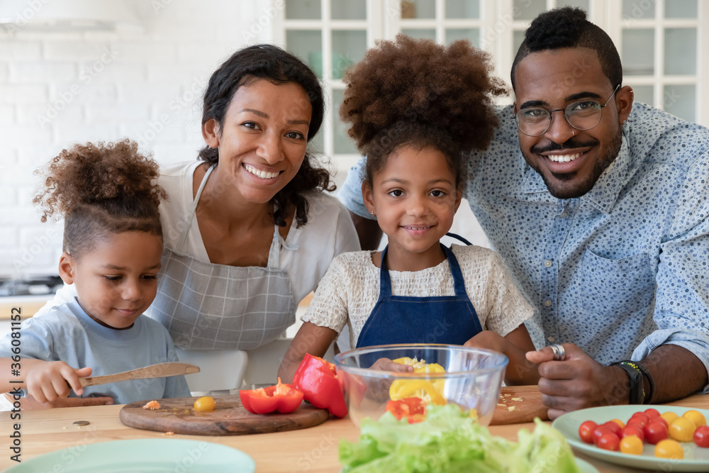 Portrait of smiling young african American family with little kids cooking together in kitchen at home, happy loving biracial parents teach small children prepare food make healthy salad dinner Portrait of smiling young african American family with little kids cooking together in kitchen at home, happy loving biracial parents teach small children prepare food make healthy salad dinner