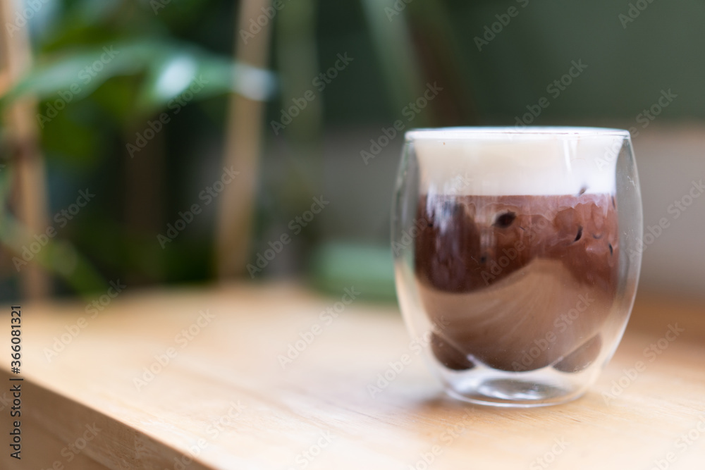 The cold chocolate top is filled with milk foam, put in 2 clear bear shaped face glasses, placed on a wooden table next to the green leafy tree in coffee shop. The cold chocolate top is filled with milk foam, put in 2 clear bear shaped face glasses, placed on a wooden table next to the green leafy tree in coffee shop.