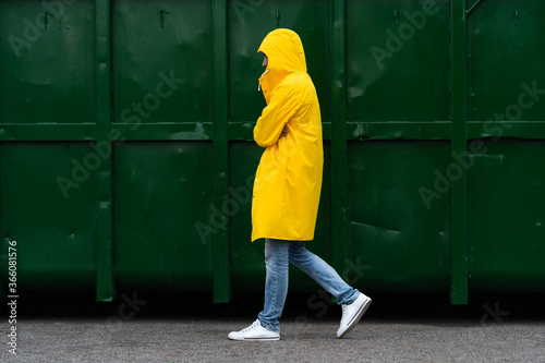 Man in a yellow raincoat walks down the street in the rain weather next to green container, side view. Outdoor. 