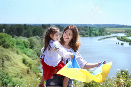 Happy smiling Ukrainian Mom and daughter in vyshyvanka (embroidered shirts) with a yellow-blue flag on a background of mountains and a river.Ukraine's Independence Day. National Flag Day of Ukraine 