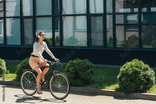 beautiful businesswoman in glasses listening music in wireless headphones and riding bicycle near building