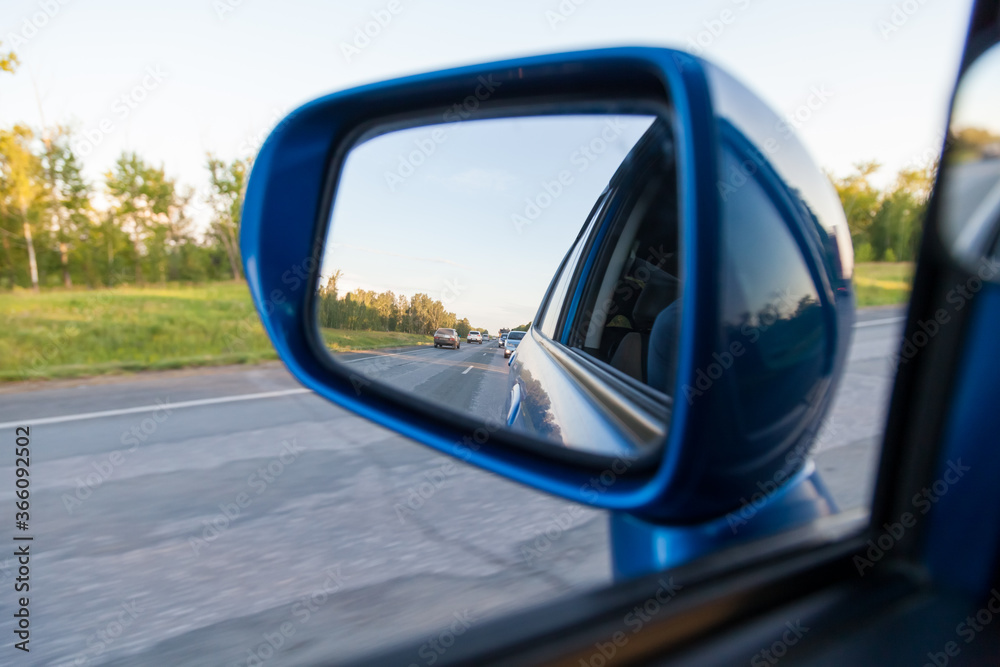 Rear view in the side mirror of a blue sedan with the reflection of heavy traffic on a country asphalt road on a summer day with green trees on the sides of the highway. Traffic Laws.