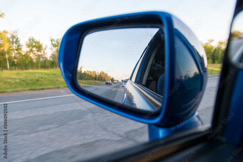 Rear view in the side mirror of a blue sedan with the reflection of heavy traffic on a country asphalt road on a summer day with green trees on the sides of the highway. Traffic Laws. Rear view in the side mirror of a blue sedan with the reflection of heavy traffic on a country asphalt road on a summer day with green trees on the sides of the highway. Traffic Laws.