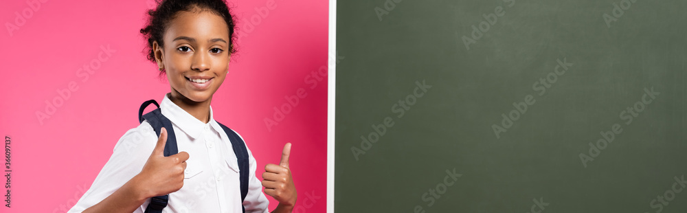 smiling african american schoolgirl with backpack near empty chalkboard showing thumbs up on pink background, panoramic shot smiling african american schoolgirl with backpack near empty chalkboard showing thumbs up on pink background, panoramic shot
