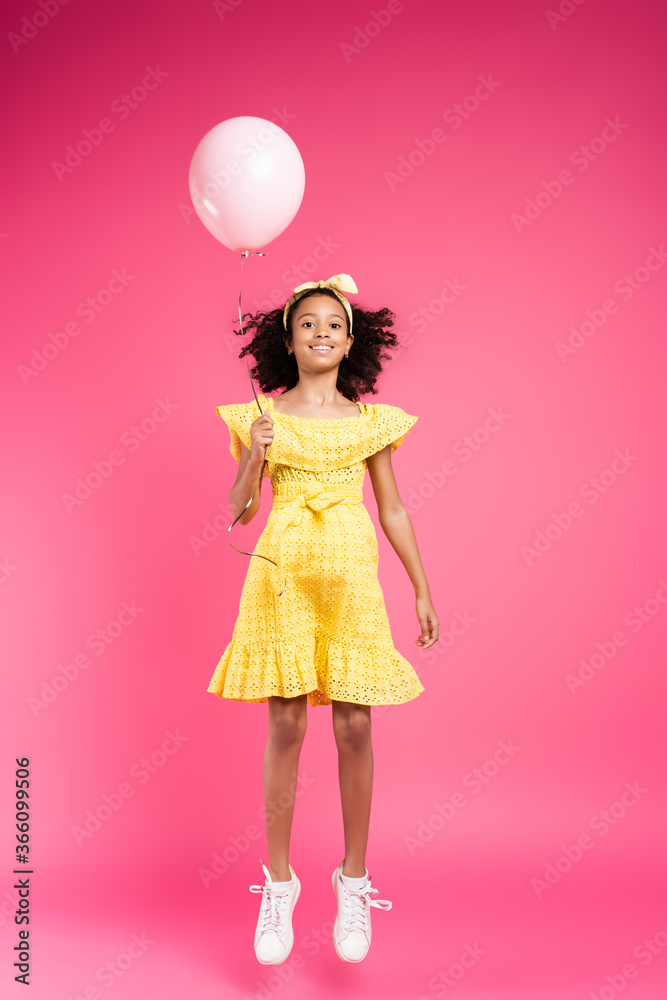 full length view of smiling curly african american child in yellow outfit flying with balloon on pink background full length view of smiling curly african american child in yellow outfit flying with balloon on pink background