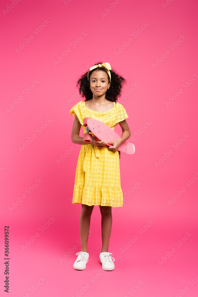 full length view of smiling curly african american child in yellow outfit with penny board on pink background