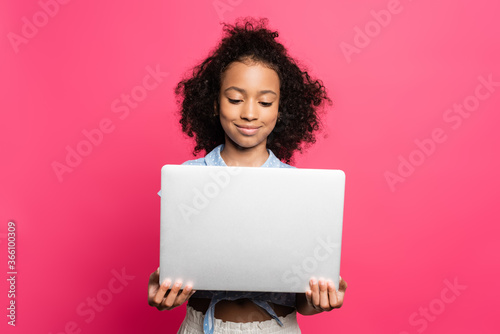 smiling cute curly african american kid holding laptop isolated on pink © LIGHTFIELD STUDIOS