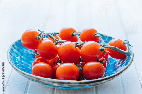Appetizing ripe cherry tomatoes on a twig in a blue clay plate on a white wooden background. Fresh and healthy vegetarian food.