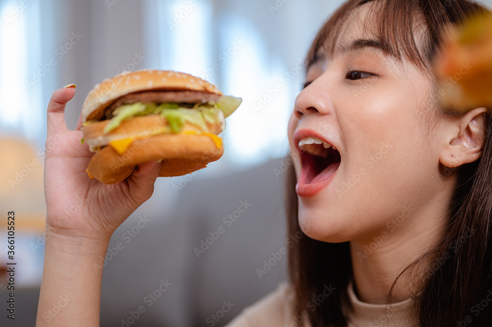 Hungry young woman eating junk food hamburger and pizza for lunch by ordering delivery at home on holiday. unhealthy meal, obesity risk. Hungry young woman eating junk food hamburger and pizza for lunch by ordering delivery at home on holiday. unhealthy meal, obesity risk.
