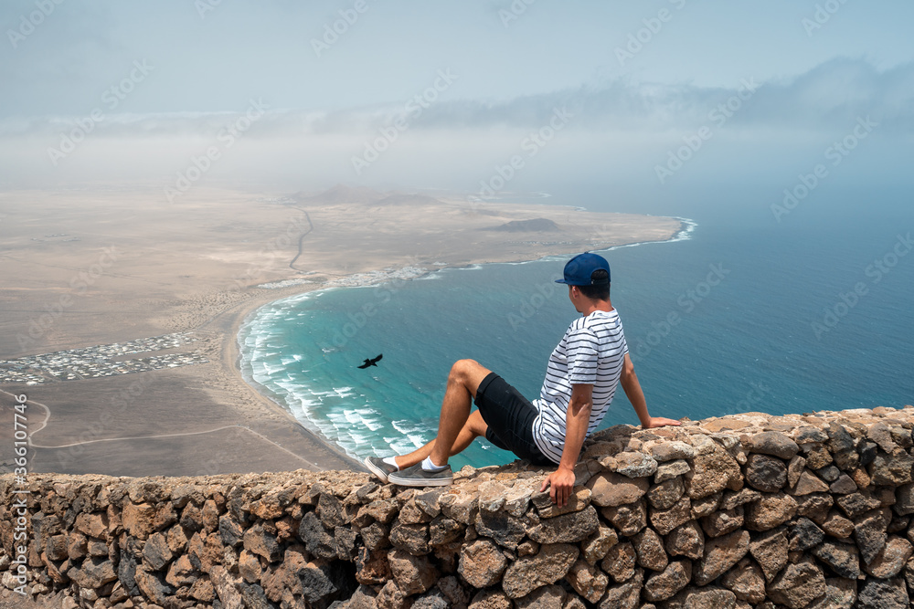 Hombre joven sentado en un muro de piedras en la cima de un acantilado observa el pueblo de Famara mientras un cuervo vuela a su lado. Teguise. Lanzarote. Islas Canarias