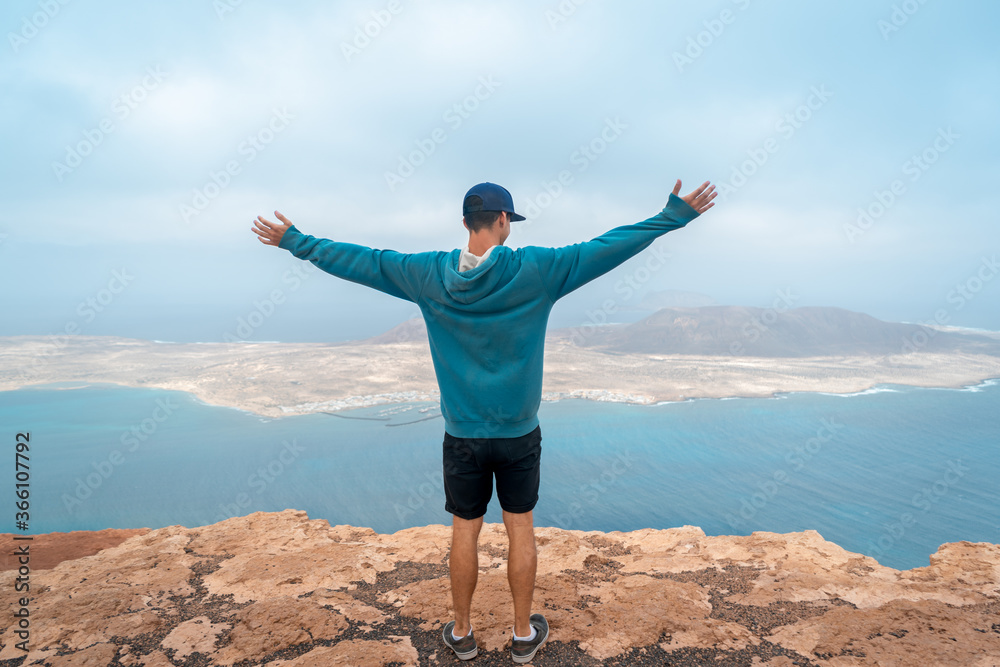 Joven viajero en la cima de un acantilado en la isla de Lanzarote, se admira con la vista de la isla de La Graciosa. Haría. Lanzarote. Islas Canarias Joven viajero en la cima de un acantilado en la isla de Lanzarote, se admira con la vista de la isla de La Graciosa. Haría. Lanzarote. Islas Canarias