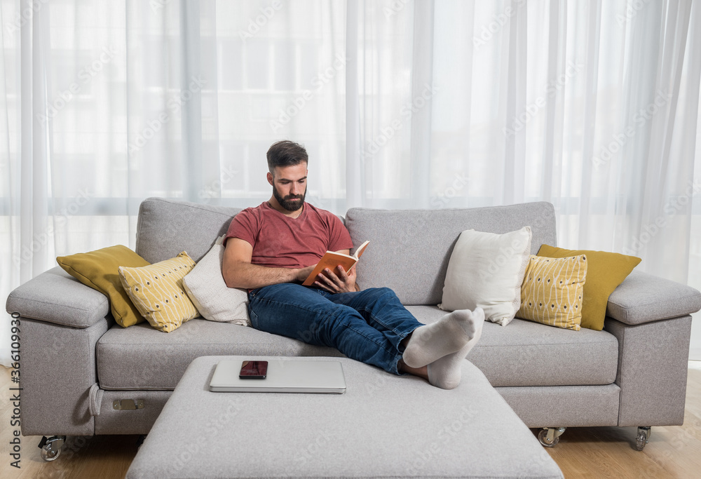 Young beautiful man or guy lying on the sofa at home reading the book and enjoying his free time without his wife and children with smile and happy face Young beautiful man or guy lying on the sofa at home reading the book and enjoying his free time without his wife and children with smile and happy face