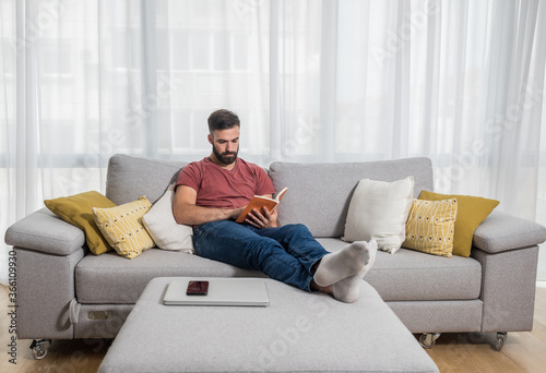 Young beautiful man or guy lying on the sofa at home reading the book and enjoying his free time without his wife and children with smile and happy face