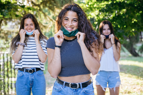Portrait of a group of young millennials friends with lowered face mask in a park at sunset at social distancing avoid the contagion from Coronavirus, Covid-19 - Beautiful women looking and smiling