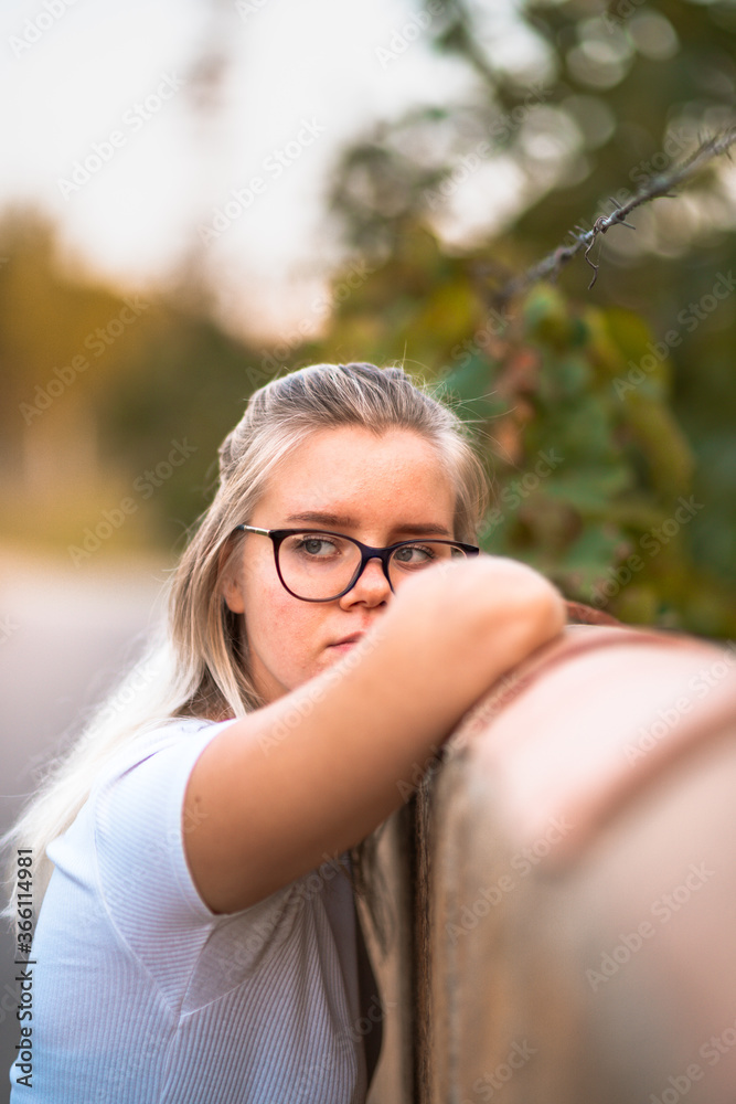 Young blonde girl resting her arm on a fence in Mallorca, Spain. Summer 2020 She is wearing a white t-shirt and black eyeglasses. Her blonde hair is half up and half down. Young blonde girl resting her arm on a fence in Mallorca, Spain. Summer 2020 She is wearing a white t-shirt and black eyeglasses. Her blonde hair is half up and half down.