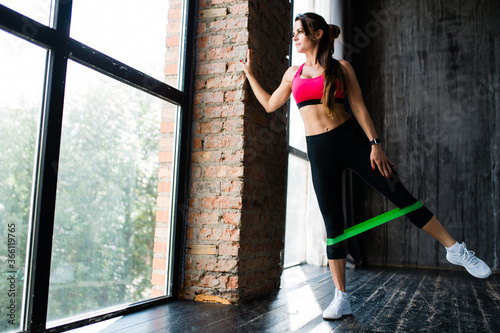 Girl doing exercises with a sports rubber band on her legs lifting her leg to the side and leaning on the wall in front of the panoramic window in the loft