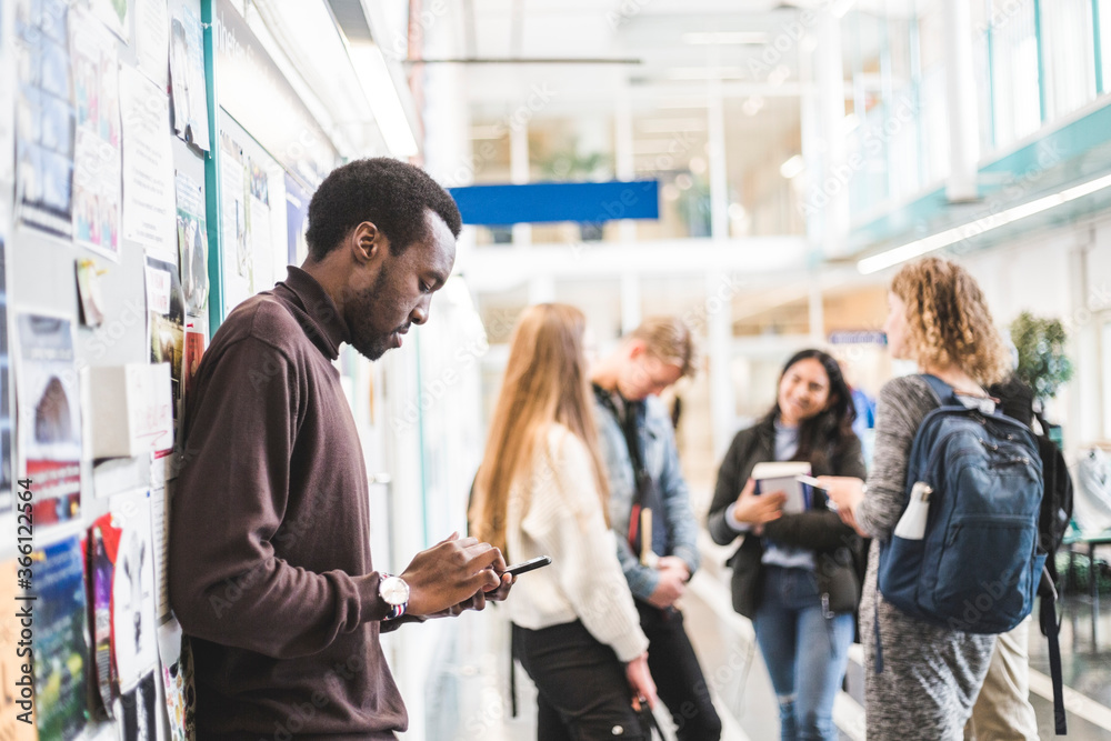 Side view of young male student using mobile phone while leaning on wall at university Side view of young male student using mobile phone while leaning on wall at university
