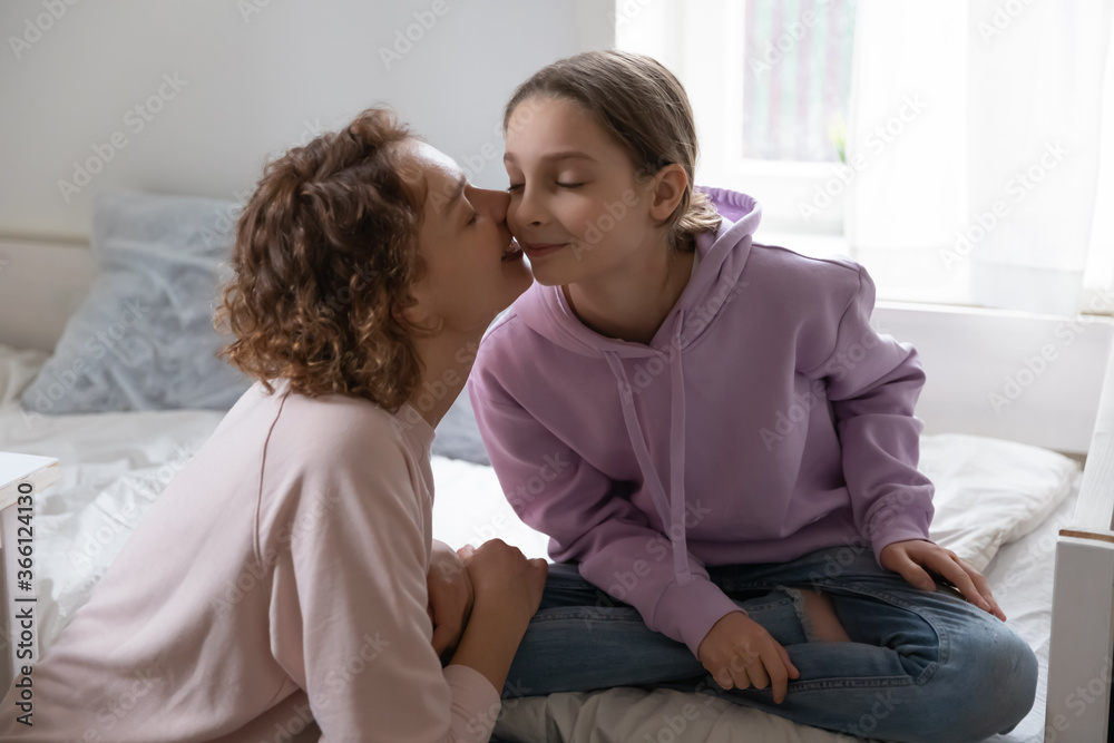 Loving mother kissing adorable teenage pretty daughter on cheek, caring overjoyed mum and happy teenager girl enjoying tender moment, sitting on bed in bedroom at home, good family relationship