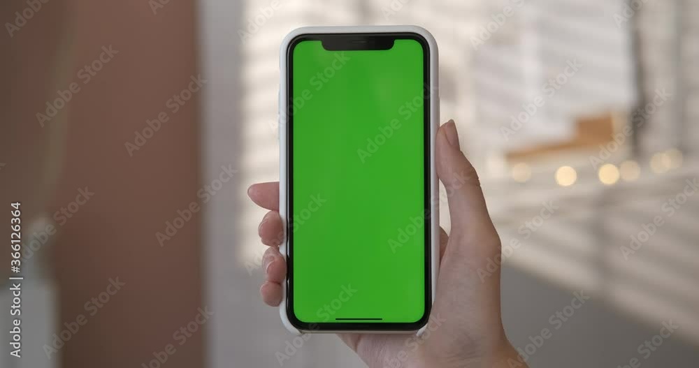 Girl using smartphone with green screen in living room watching movie, video content. Close up woman's hand holding smart phone in a vertical position. Indoors. Touch screen hand gestures. Closeup