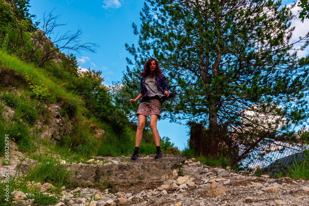 Fototapeta premium A full body shot of an energetic young Caucasian redhead woman jumping on a hiking path in the mountains under warm summer sunlight (Puget-Theniers, France)