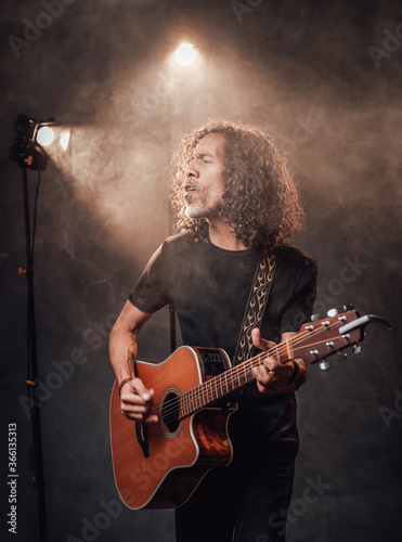 Hispanic musician in black t-shirt emotionally singing and playing guitar in stage lights, surrounded by smoke © Fxquadro