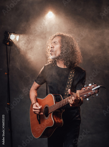 Hispanic musician in black t-shirt emotionally singing and playing guitar in stage lights, surrounded by smoke