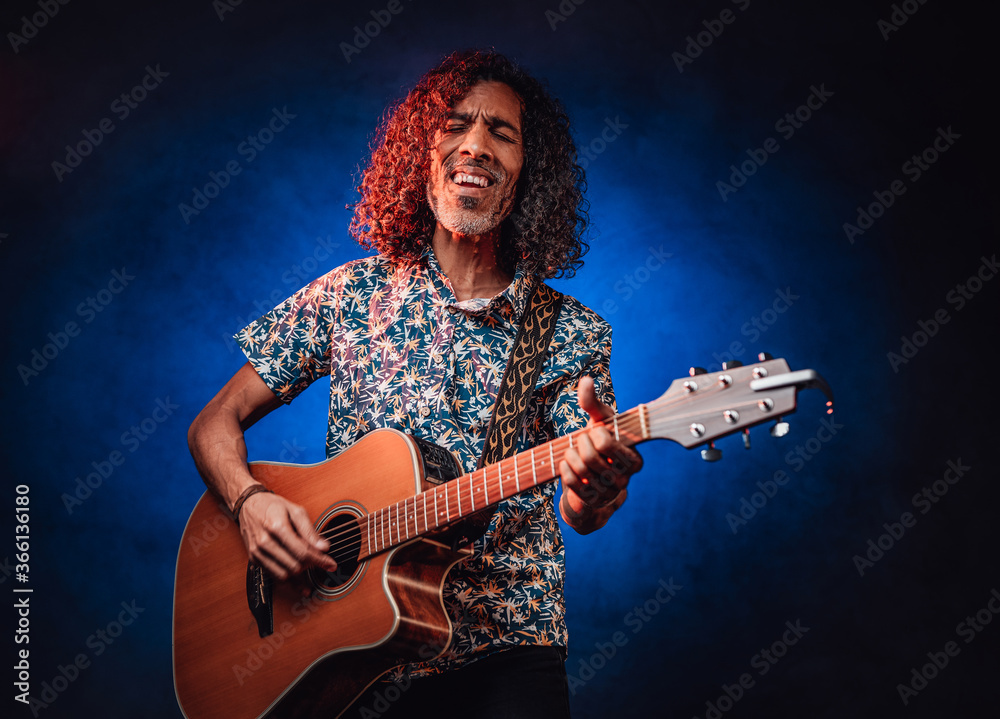 Obraz premium Hispanic musician emotionally singing and playing guitar on a dark lluminated by blue and red light. Concept of music, hobby, festival