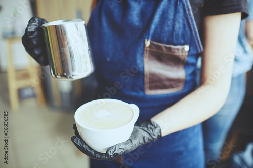 Female holds a milk jug and a cup of coffee, makes cappuccino, pouring whipped milk into espresso.Blurred image, selective focus