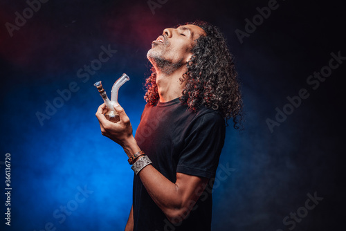 Middle aged hispanic male with long curly hair smoking from bong on a dark background illuminated by blue light