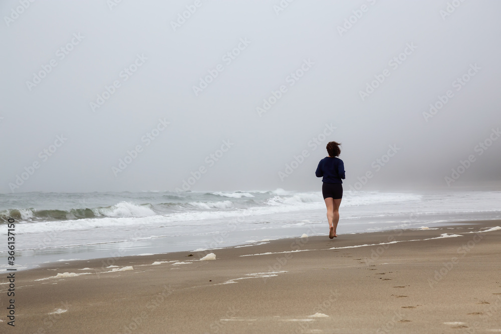 Athletic Caucasian Girl Running on a Sandy Beach near Pacific Ocean Coast during a cloudy and foggy morning. Taken at Raft Cove, Vancouver Island, British Columbia, Canada. Athletic Caucasian Girl Running on a Sandy Beach near Pacific Ocean Coast during a cloudy and foggy morning. Taken at Raft Cove, Vancouver Island, British Columbia, Canada.