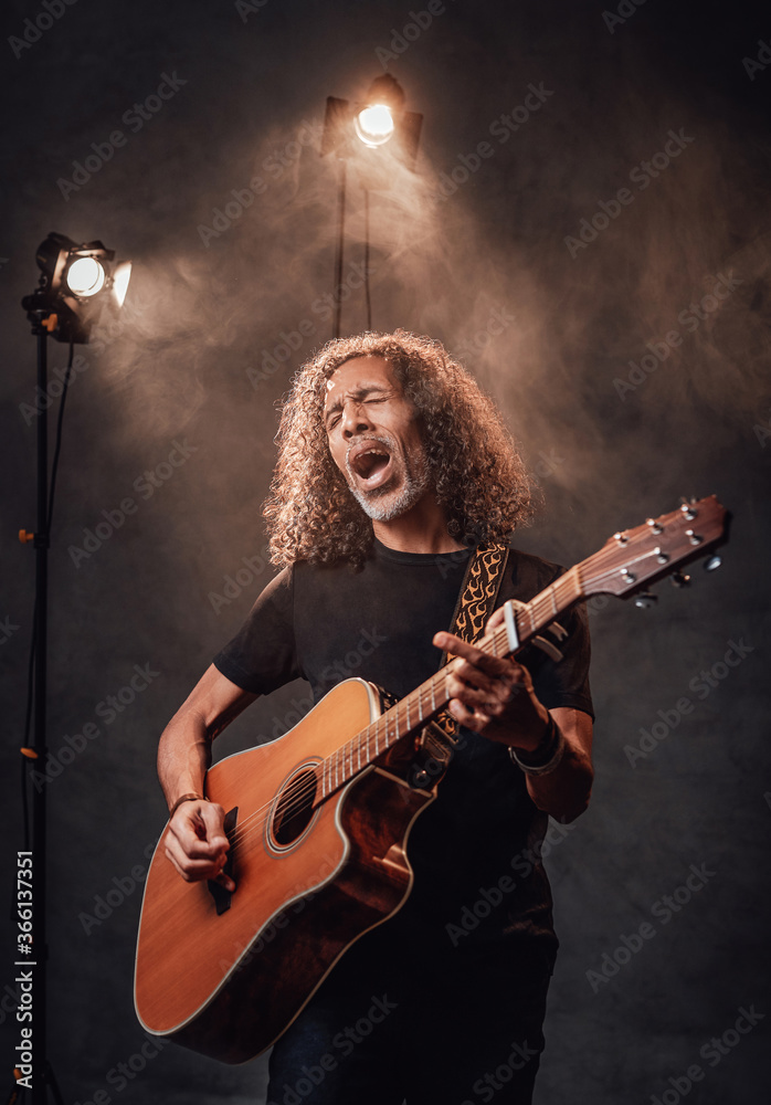 Middle aged hispanic musician in black t-shirt emotionally singing and playing guitar. View of musician in the spotlight