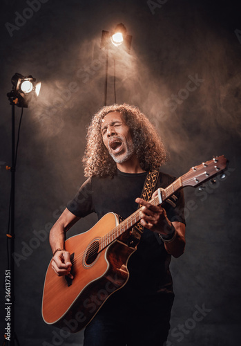 Middle aged hispanic musician in black t-shirt emotionally singing and playing guitar. View of musician in the spotlight