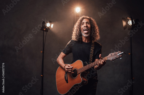 Middle aged hispanic musician in black t-shirt emotionally singing and playing guitar. View of musician in the spotlight