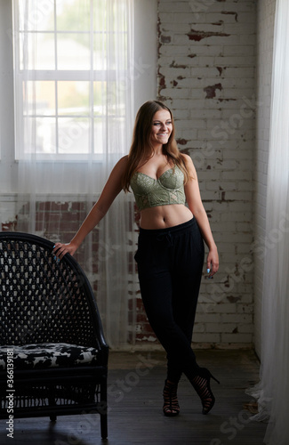 Stunning young woman wearing a green bustier top and black pants stands next to a black wicker chair - studio fashion