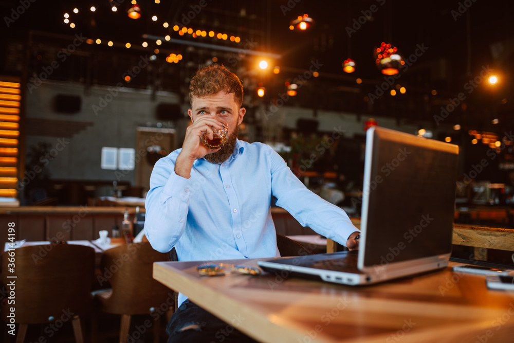 A young handsome caucasian businessman with a beard is sitting in a cafe drinking whiskey and working on a laptop. Work in a restaurant