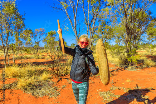 Happy tourist woman holding an aboriginal weapon of spear and a wooden shield used by Luritja and Pertame people, Central Australia, Northern Territory. Red sand and bush wilderness in desert outback.